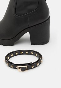 Botine negre cu toc, din piele texturată, cu o talpă masivă, însoțite de un curea neagră asortată, decorată cu prefețe aurii și o cataramă.