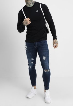 Schwarzes langärmliges Nike-Shirt, abgenutzte blaue Jeans mit Rissen und weiße Sneaker. Merkmale tätowierte Arme und Riemen am Shirt.