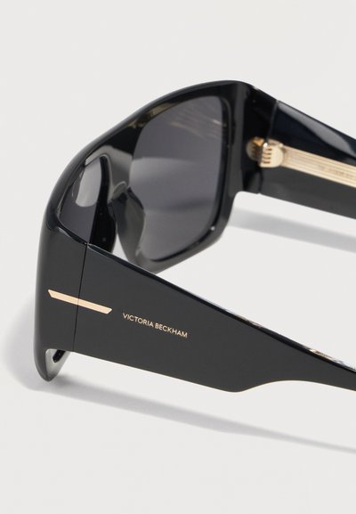 Μαύρα υπερμεγέθη γυαλιά ηλίου με χοντρά γυαλιστερά πλαίσια και χρυσές λεπτομέρειες, με το λογότυπο "Victoria Beckham" στον βραχίονα.