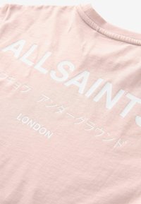 Světle růžové bavlněné tričko s kulatým výstřihem; obsahuje velký bílý text: "ALLSAINTS" a "LONDÝN" spolu s japonskými znaky.