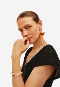 Zlatý �řetízkový náhrdelník, růžový korálkový náramek, velké náušnice s oranžovou květinou; model v texturovaném černém topu, ruka na bradě, neutrální pozadí.