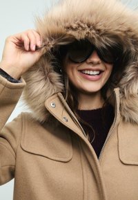 Cappotto in misto lana beige con cappuccio in pelliccia sintetica removibile, chiusura con zip, finiture in metallo argentato e due tasche sul petto. Indossato sopra un maglione scuro.