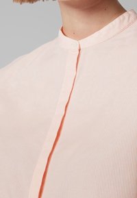 Blouse rayée rose clair avec un col mandarin, dotée d'une fermeture à bouton unique et d'une texture subtile dans le tissu.