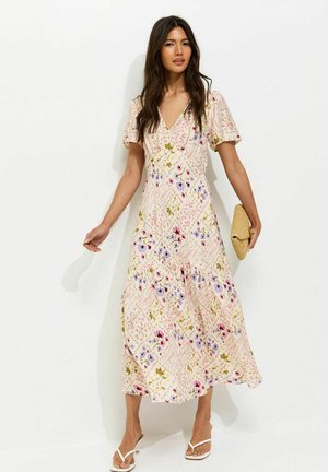 Femme portant une robe midi à fleurs avec des manches courtes bouffantes, tenant une pochette tissée et portant des sandales à talons blanches sur un fond uni.