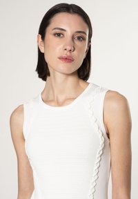 Robe blanche sans manches avec une texture côtelée et des volants décoratifs le long des coutures latérales, mettant en valeur une silhouette ajustée.