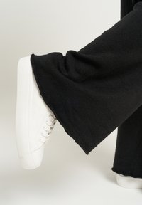 Białe sneakersy o teksturowanym wykończeniu, z wiązaniami i grubą podeszwą. Czarne spodnie dzwony z miękkiej, tkanej tekstury, częściowo zakrywające buty.