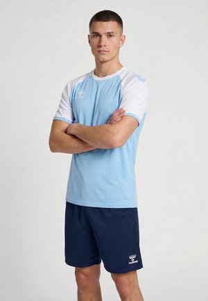 Ung mand stående med armene krydset, iført lyseblå og hvid sportstrøje og mørkeblå shorts mod en ensfarvet baggrund.