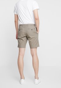 Mann in beige knielangen Shorts, weißem T-Shirt und weißen Sneakers, steht mit den Händen in den Taschen und schaut weg.