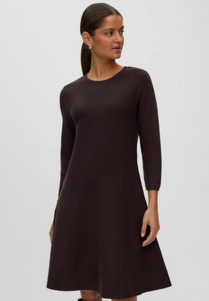 Femme aux cheveux foncés attachés, portant une robe en maille marron foncé, à manches longues et longueur genou, sur un fond clair uni.