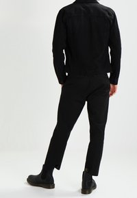 Zwarte denimjack met een gestructureerd ontwerp, weergegeven vanaf de achterkant. Gecombineerd met slim-fit zwarte broeken en zwarte enkellaarsjes.