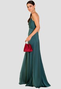 Lungo vestito di raso verde con spalline sottili, silhouette fluida e profondo scollo sulla schiena. Presenta una piccola borsa in raso rosso con manico di perline.