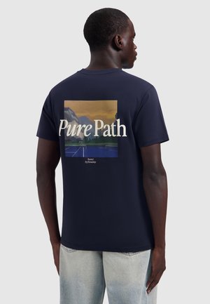 Mann trägt ein marineblaues T-Shirt mit Berg- und Tennisplatzgrafik und dem Text "Pure Path" auf dem Rücken, kombiniert mit hellblauen Jeans.