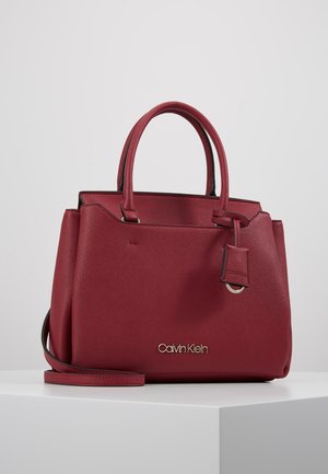 Sac à main rouge Calvin Klein avec deux poignées, une bandoulière amovible et une étiquette de marque suspendue sur le côté.
