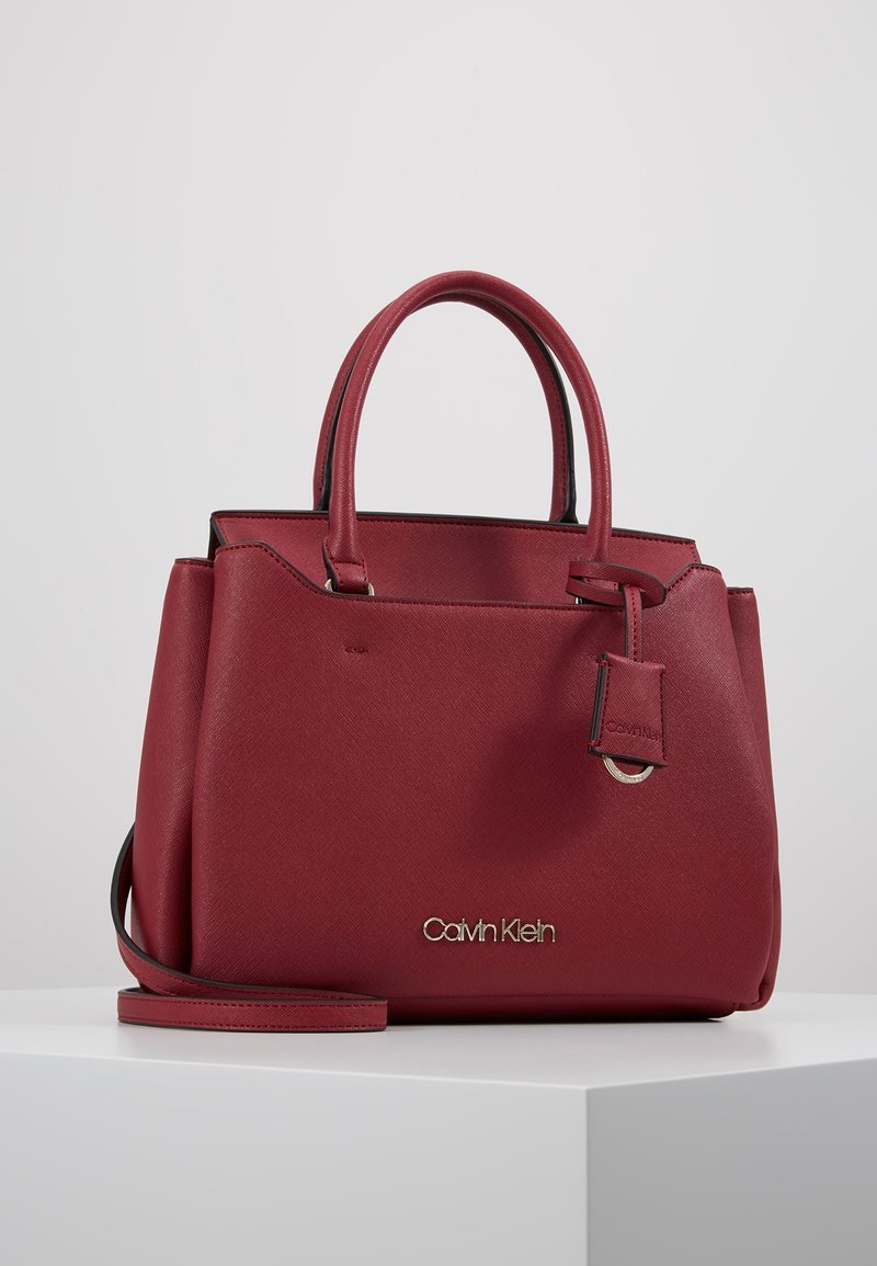 Sac à main rouge Calvin Klein avec deux poignées, une bandoulière amovible et une étiquette de marque suspendue sur le côté.