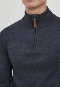 Dunkelgrauer Strickpullover mit hohem Kragen und kurzem Reißverschluss am Hals. Beinhaltet ein Lederakzent in der Nähe des Reißverschlusses für zusätzliche Details.