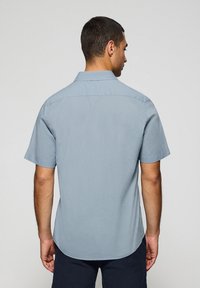 Camicia a maniche corte in tessuto blu chiaro, con colletto standard, piega sul retro e orlo curvo. Texture liscia senza motivi visibili.