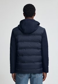 Mörkblå vadderad jacka med huva, med horisontella quiltade sektioner och elastiska ärmslut. Slät textur och minimalistisk design.