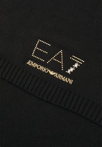 Zwarte gebreide stof met geribbelde zoom, versierd met goudkleurige pailletten tekst "EA7" en het "Emporio Armani" logo. Glad textuur met decoratieve accenten.