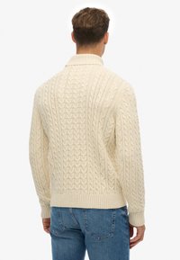 Mand iført en cremet kabelstrikket sweater og blå jeans, stående med ryggen til mod en enkel hvid baggrund.