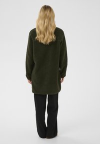 Zielono-oliwkowa bluza polarowa z długimi rękawami, lekko oversize'owym krojem, teksturowaną powierzchnią i zaokrąglonym dołem, w połączeniu z czarnymi szerokimi spodniami.