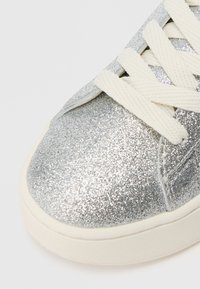 Baskets argentées scintillantes avec une tige texturée, des lacets blancs doux et une semelle blanche lisse. Bout arrondi et détails de couture discrets.