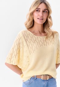 Lichtgele gebreide trui met een losse pasvorm, voorzien van een golvenpatroon en korte mouwen. Gecombineerd met hooggesneden blauwe jeans en een beige riem.