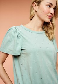 Mintgrøn t-shirt med korte puffærmer og spredte rhinsten-dekorationer. Blødt stof og afslappet pasform.