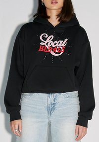 Czarny krótki hoodie z przednią kieszenią, białym i czerwonym napisem "Local Heroes" z dekoracyjnymi akcentami oraz miękką teksturą.