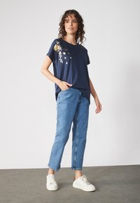 Navy blue short-sleeve T-shirt with gold floral embroidery, paired with light blue denim jeans and white sneakers. Features side pockets.