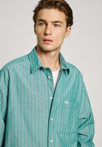 Camicia a righe verde chiaro realizzata in un tessuto morbido. Presenta una chiusura con bottoni, una tasca sul petto e un colletto classico.