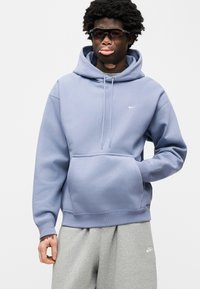 Ανοιχτό μπλε φούτερ με κουκούλα, τσέπη μπροστά, κορδόνι στην κουκούλα και μικρό λευκό λογότυπο στο στήθος. Συνδυασμένο με γκρι φόρμες.