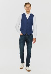 Granatowa kamizelka z sześcioma guzikami, kołnierzykiem, i kieszeniami z przodu, noszona na białej koszuli, zestawiona z ciemnymi dżinsami i białymi trampkami.