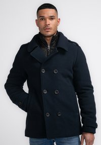 Pabellón de lana en mezcla de color navy con un frente de doble botonadura, grandes botones y amplias solapas, superpuesto sobre una chaqueta negra con cremallera.