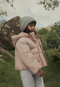 Padded rosa Jacke mit Kapuze, ausgestattet mit Taschen vorne und einem Reißverschluss, kombiniert mit einer grauen Strickmütze, getragen über grauen Sweatpants.