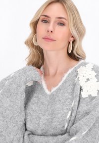 Grau gestrickter Pullover mit V-Ausschnitt, der flauschige weiße Blumenakzente an den Schultern und strukturierte Ärmel mit dekorativen Nähten aufweist.