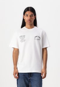 White cotton T-shirt with short sleeves. Features black text graphics on the front, including "TOMMY JEANS" and city names. Classic fit.