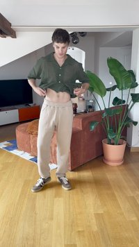 Mladý muž zvedá tmavě zelené tričko, aby odhalil břicho; stojí v obývacím pokoji s hnědou pohovkou, velkou rostlinou v terakotovém květináči a dřevěnou podlahou.