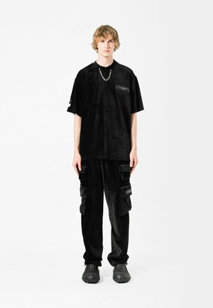 Camisa de cortadura negra de manga corta y oversized con detalle de bolsillo, combinada con pantalones cargo a juego que presentan múltiples bolsillos y calzado negro texturizado.