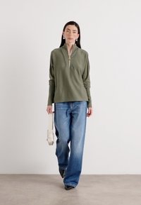 Eine Frau trägt ein olivgrünes Oberteil und blaue Jeans, hält eine hellfarbene Tasche und geht vor einer schlichten weißen Wand.