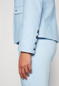 Blazer bleu clair avec une texture lisse, un design structuré et cinq boutons noirs en décoration sur la manche. Coupe ajustée.