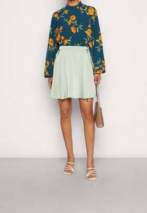 Bloemrijke top met lange mouwen in donker teal met oranje bloemen, gecombineerd met een licht muntgroene flared rok. Het model houdt een gestructureerde bruine handtas vast.