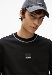 Fekete kapucnis pulóver kontraszt varrással, négyzet alakú logóval. Szürke sapka lapos karimával, texturált anyagból.