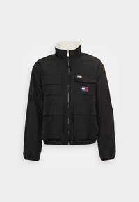 Czarna, lekka kurtka z wysokim kołnierzem, zapinana na zamek z przodu, z wieloma przednimi kieszeniami. Posiada naszywkę z logo oraz subtelną, teksturowaną tkaninę.