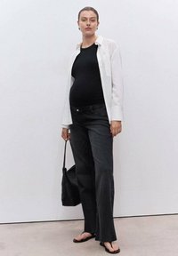 Pregnant woman wearing a black top, white unbuttoned shirt, black jeans, flip-flops, holding a black woven handbag against a plain wall.