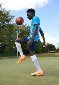 Ghete de fotbal portocalii și roz strălucitor, tricou grafic albastru, pantaloni scurți închisi la culoare și șosete albe; jonglând cu o minge de fotbal neagră și portocalie pe un teren.