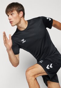 Camiseta deportiva negra y pantalones cortos, hechos de un tejido ligero. Presenta un logo en el pecho y un diseño geométrico en los pantalones cortos.