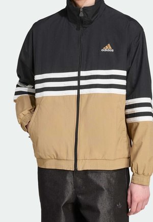 Mężczyzna w czarno-beżowej kurtce sportowej Adidas z białymi paskami i czarnych spodniach, ręce w kieszeniach.