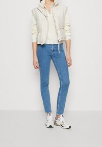 Lehká béžová prošívaná vesta přes krémový svetr, spárovaná s přiléhavými modrými džínami a bílými teniskami s černými a béžovými akcenty.