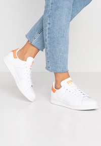 White sneakers with orange accents, featuring a smooth leather upper, rounded toe, and perforated detailing for ventilation.