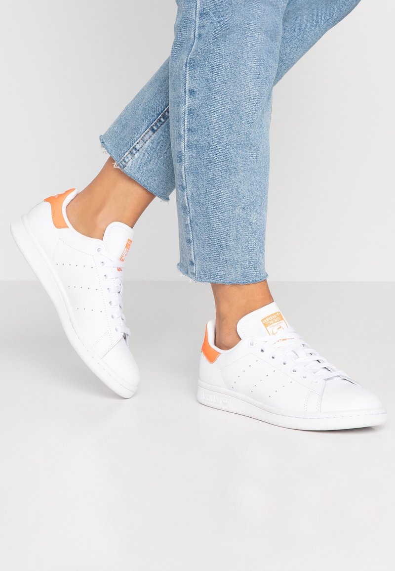 White sneakers with orange accents, featuring a smooth leather upper, rounded toe, and perforated detailing for ventilation.